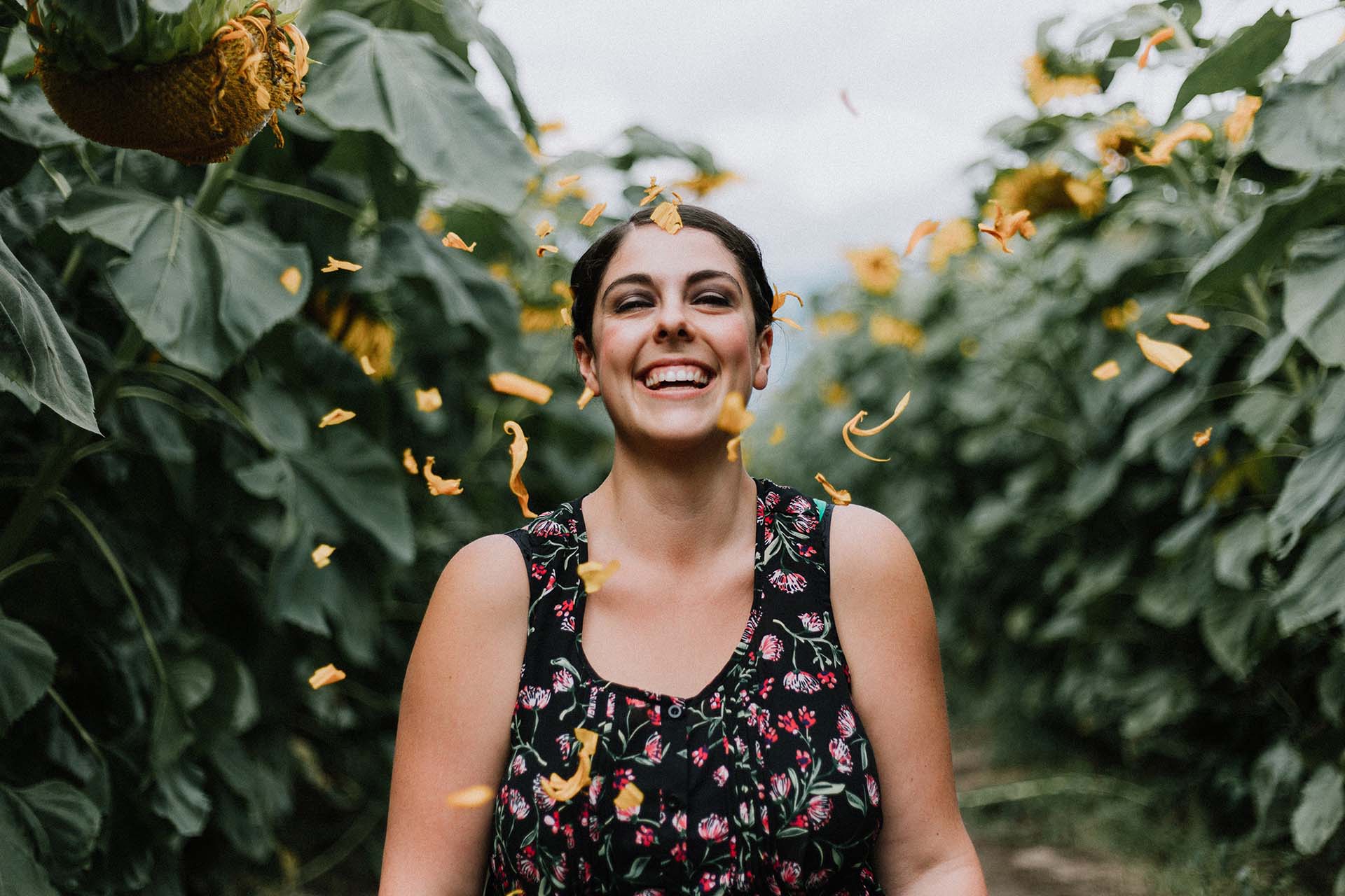 Young happy woman with vitality throwing yellow sunflower petals in the air with vitality in a sunflower garden